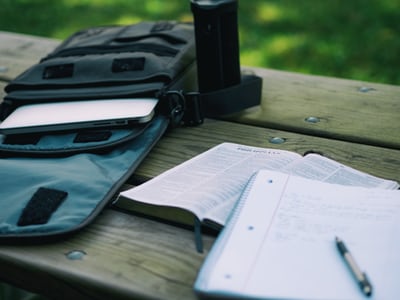 A work bag lies open next to an open book with a notebook and pen on top.